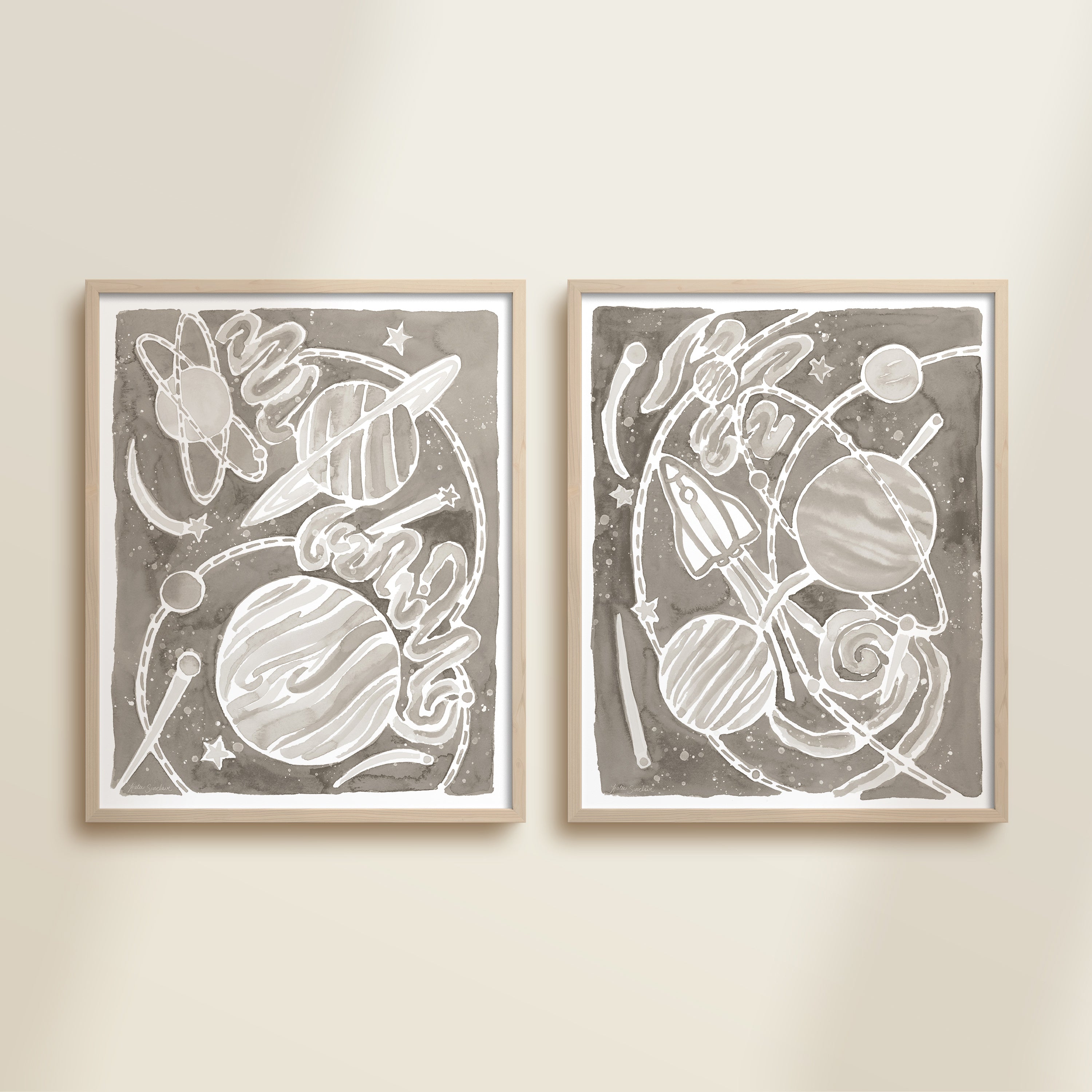 Two framed gray watercolor space-themed artworks on a beige wall, featuring planets, spaceships, stars, and a galaxy.