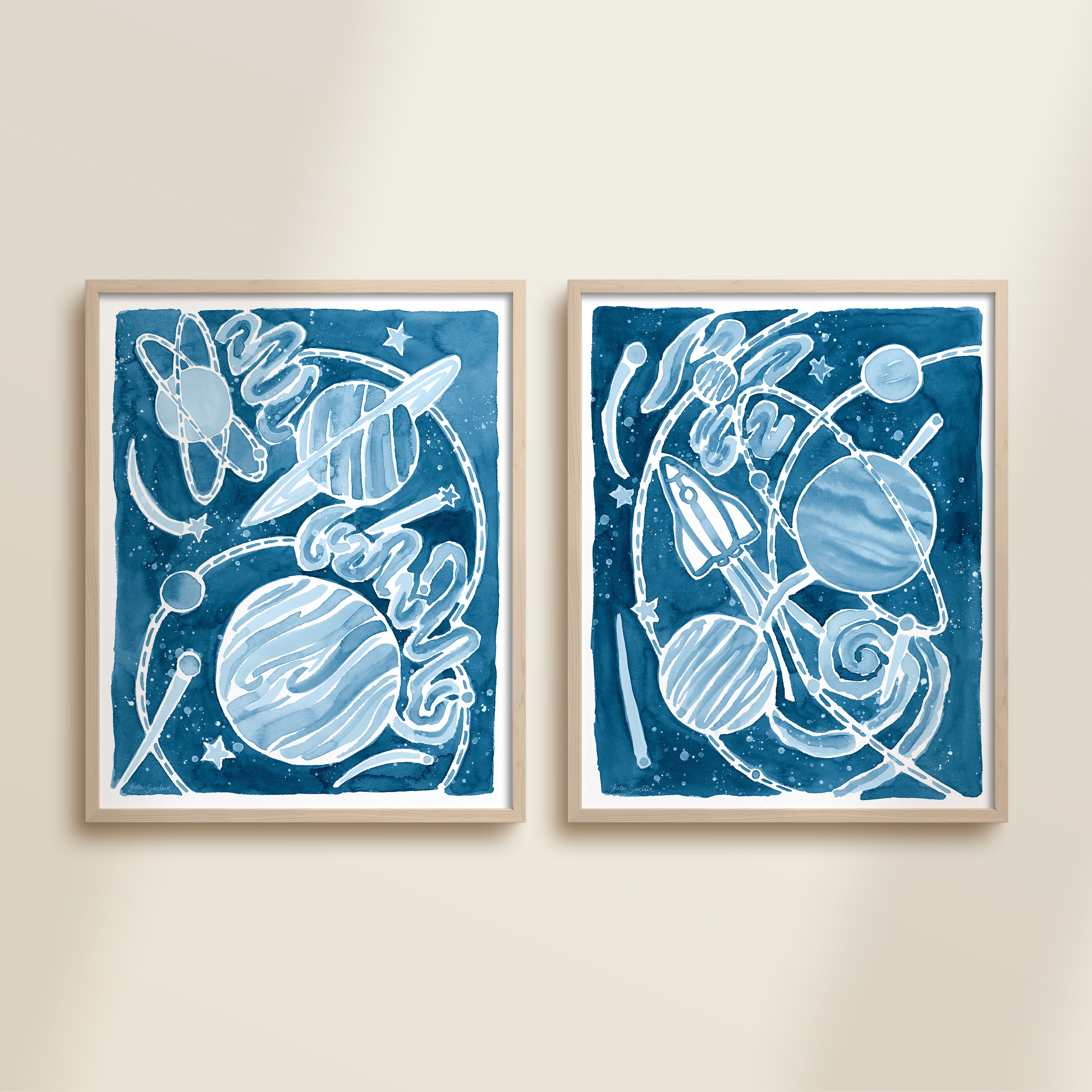 Two framed blue watercolor space-themed artworks on a beige wall, featuring planets, spaceships, stars, and a galaxy.