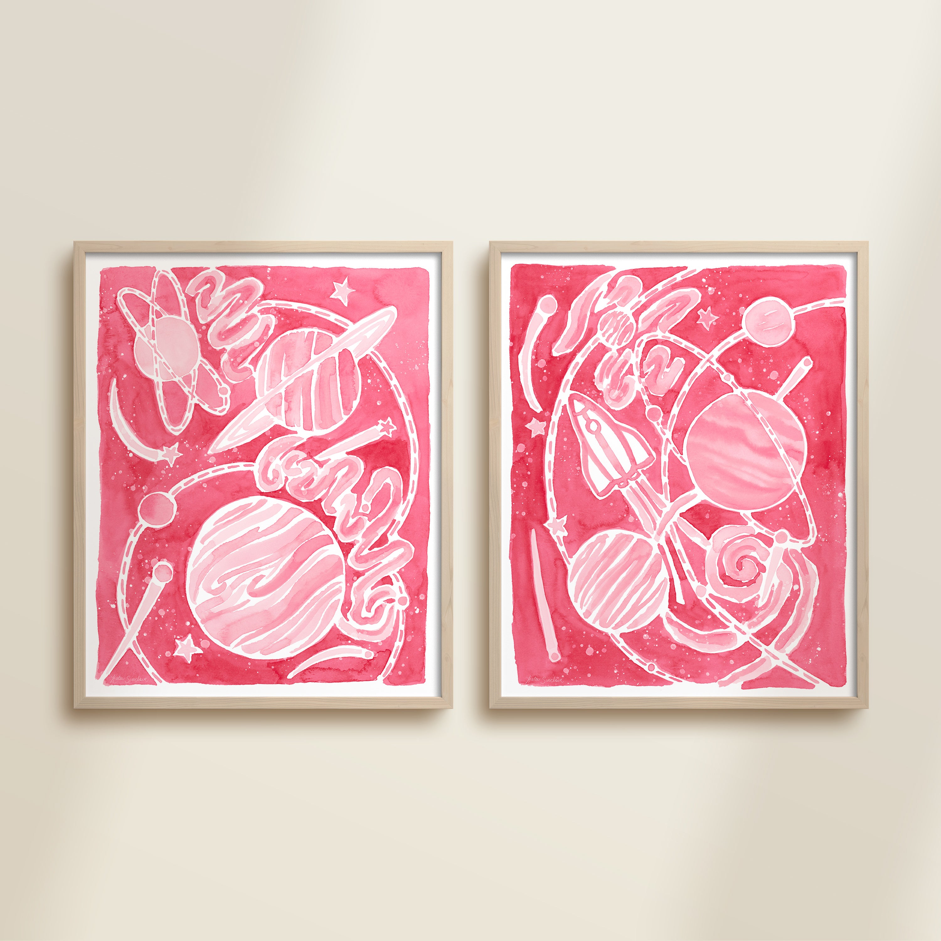 Two framed pink watercolor space-themed artworks on a beige wall, featuring planets, spaceships, stars, and a galaxy.