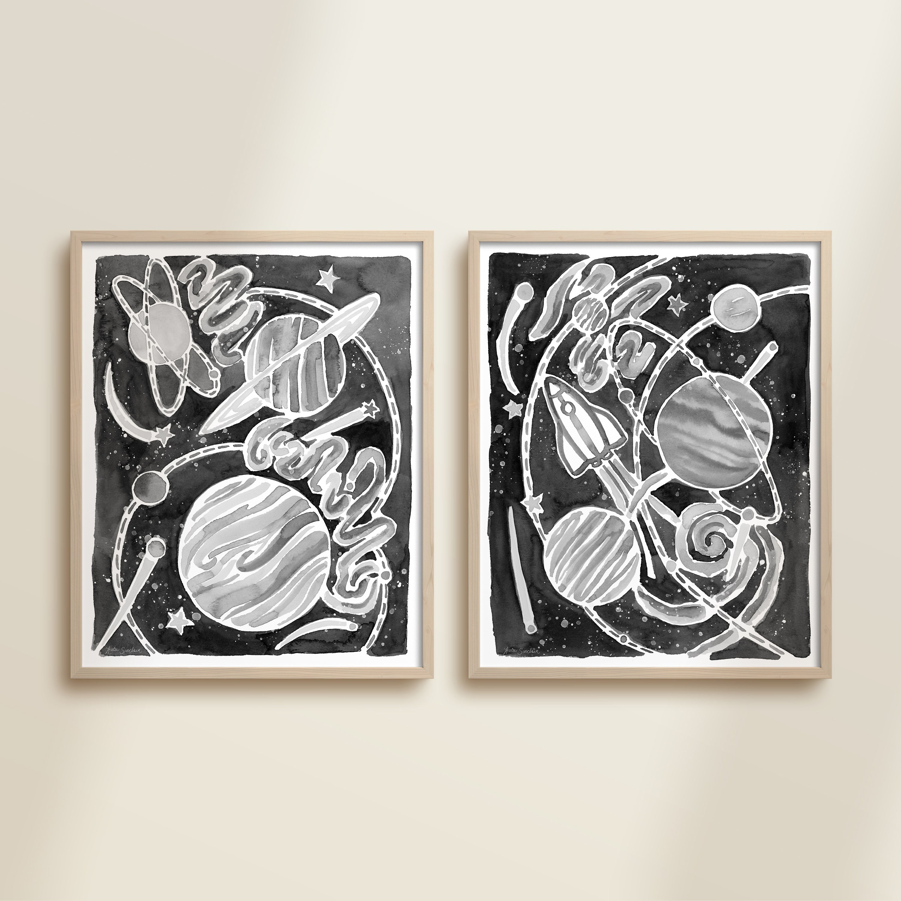 Two framed black and white space-themed artworks on a beige wall, featuring planets, spaceships, stars, and a galaxy.