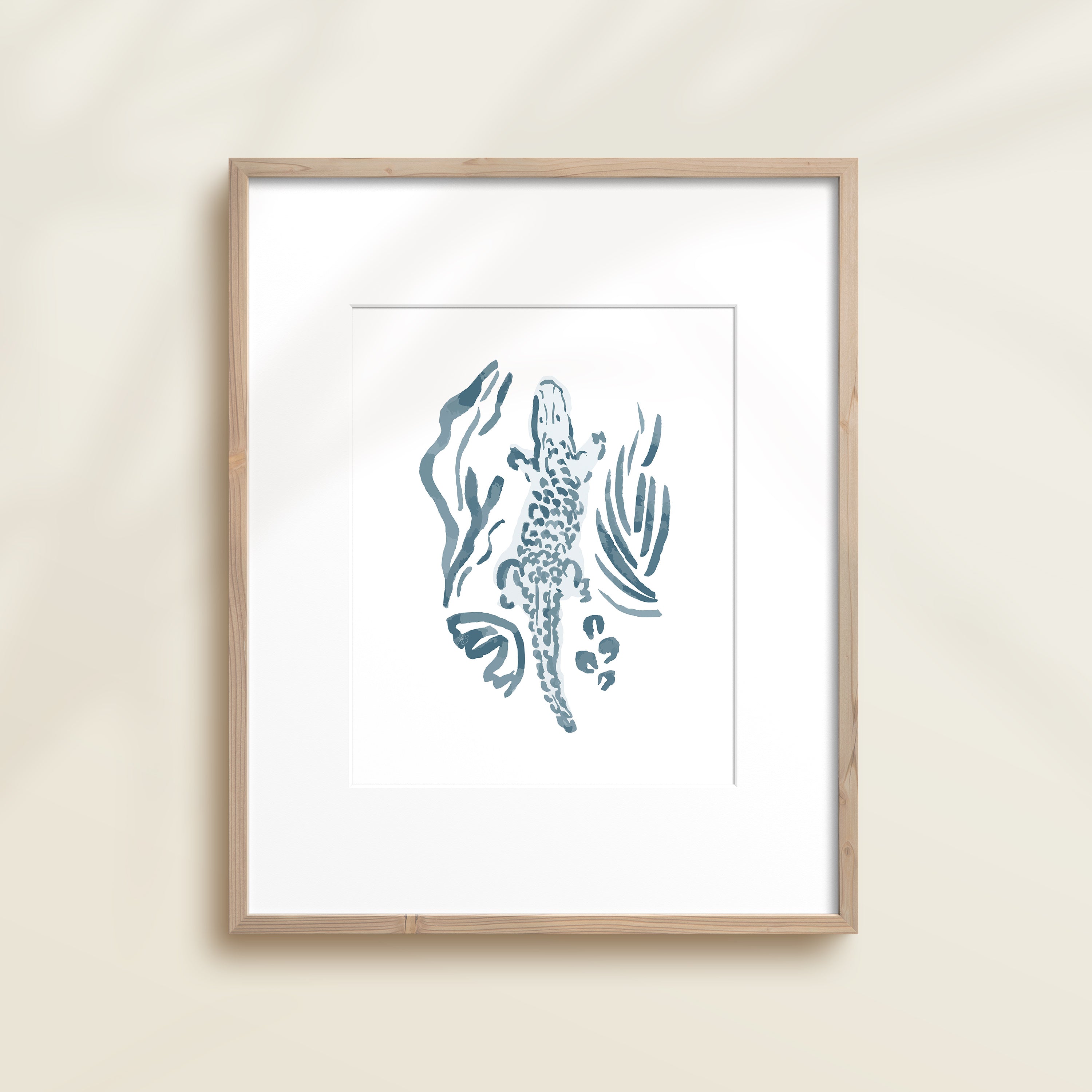 A framed watercolor print by Katie Sinclair of a crocodile surrounded by  foliage, displayed on a wall.  This image showcases the blue color version.