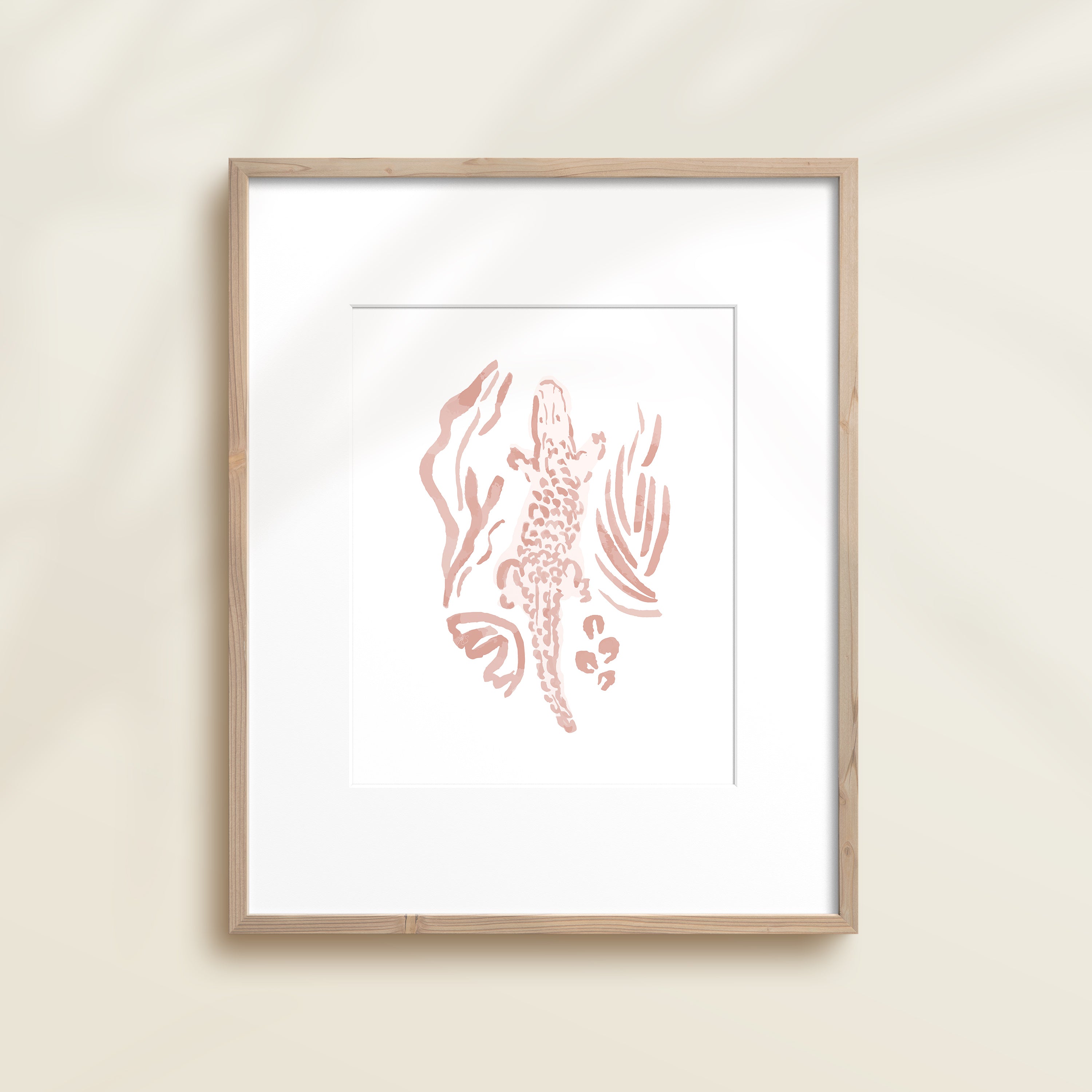 A framed watercolor print by Katie Sinclair of a crocodile surrounded by  foliage, displayed on a wall.  This image showcases the pink color version.