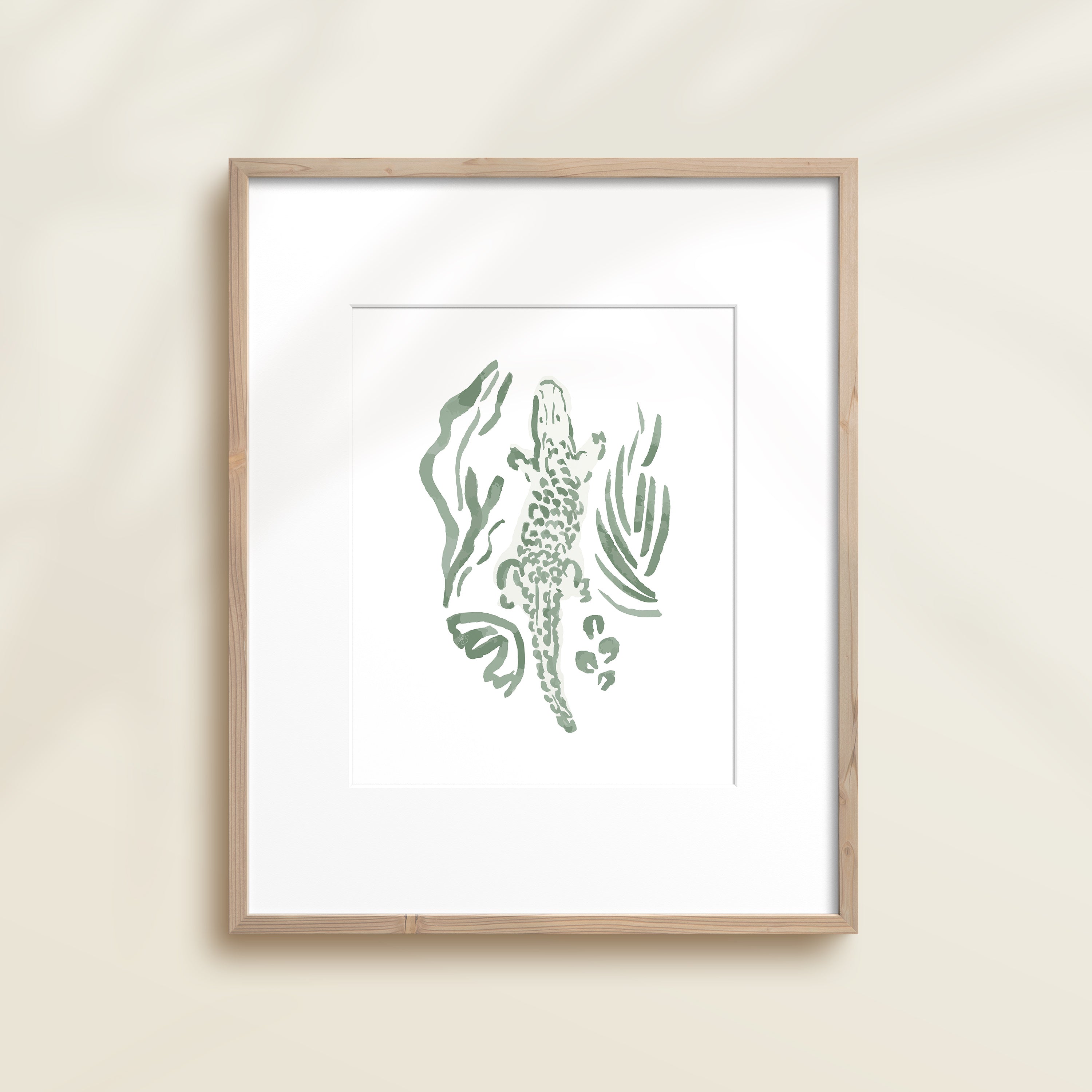 A framed watercolor print by Katie Sinclair of an crocodile surrounded by  foliage, displayed on a wall.  This image showcases the green color version.