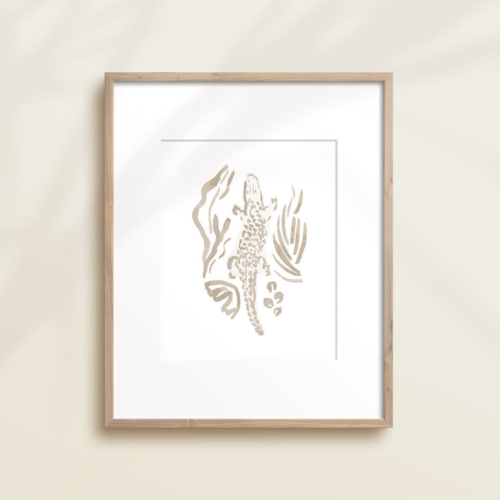 A framed watercolor print by Katie Sinclair of an crocodile surrounded by  foliage, displayed on a wall.  This image showcases the gray color version.