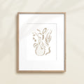 A framed watercolor print by Katie Sinclair of an sloth surrounded by  foliage, displayed on a wall.  This image showcases the gray color version.