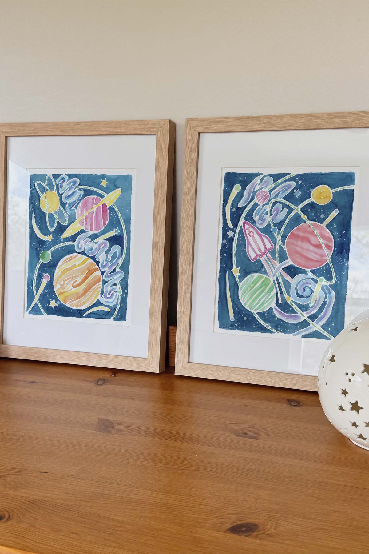 Space Explorer and Through the Galaxy are a colorful watercolor art print set featuring outer space, stars, galaxies, planets, and spaceships. Perfect for kids’ rooms, nurseries, playrooms.  It shows a framed archival print set in wooden frames on a dresser.