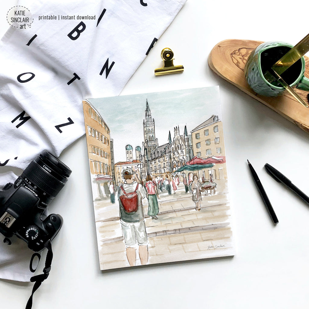 Flat lay of a drawing depicting a person taking a photo in a European square with historic buildings. Surrounding items include a camera, pens, a cup with brushes, a white cloth with black letters, a gold clip, and a wooden coaster.